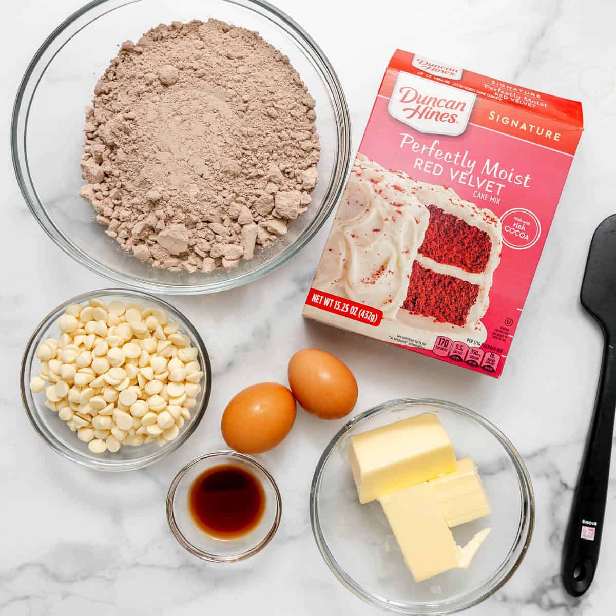 Ingredients for red velvet cake: a box of cake mix, white chocolate chips, two eggs, vanilla extract, and sticks of butter in glass bowls, with a black spatula on a marble surface.