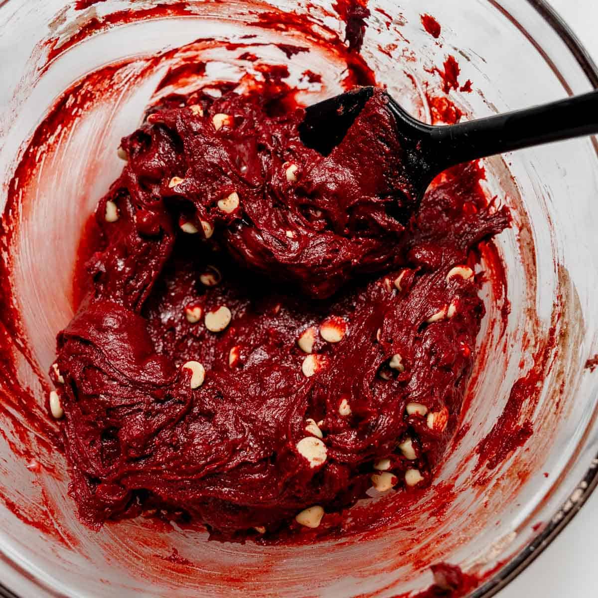 Red velvet cookie dough with white chocolate chips in a glass mixing bowl with a black spatula.