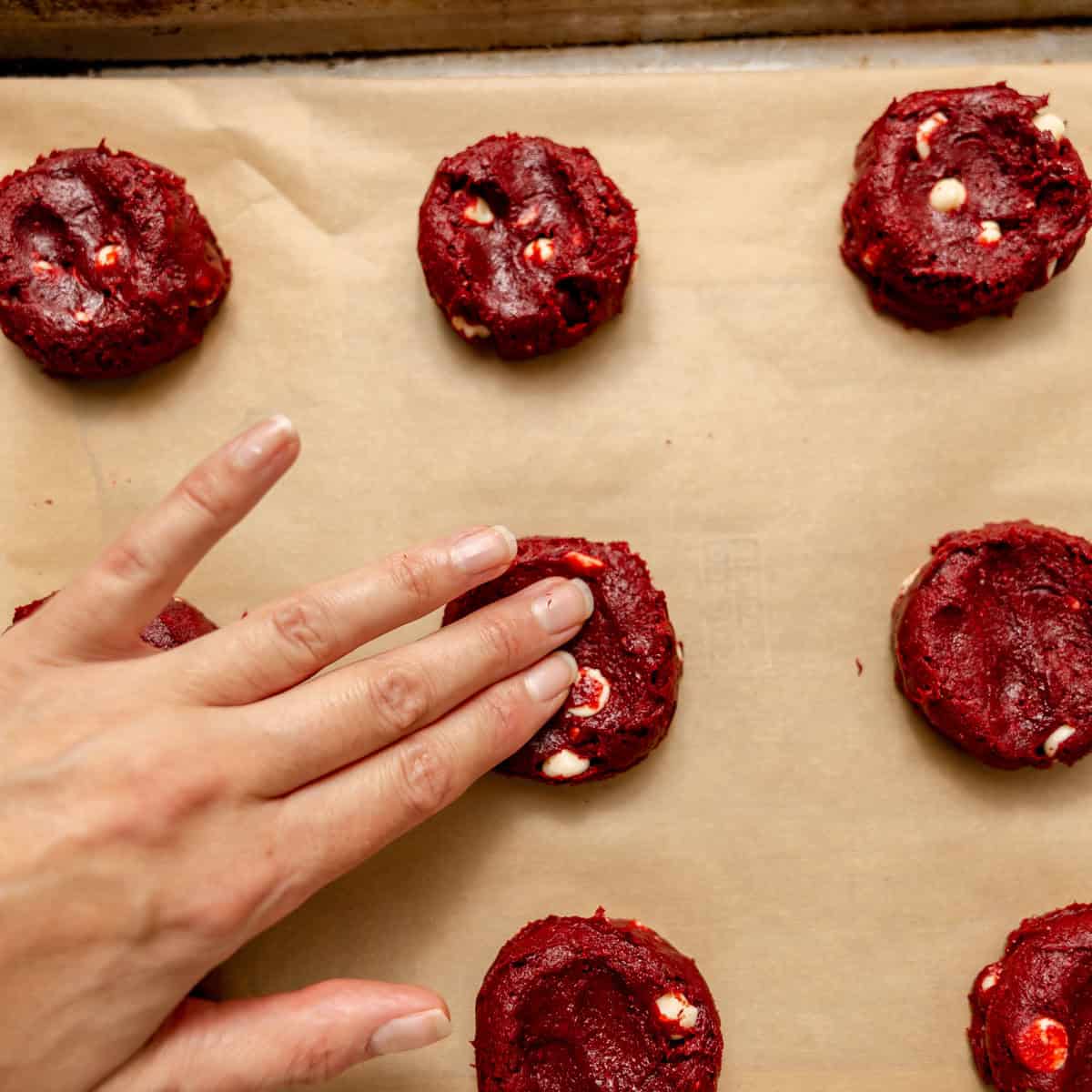 A hand presses down on a raw red velvet cookie dough ball on a parchment-lined baking sheet.