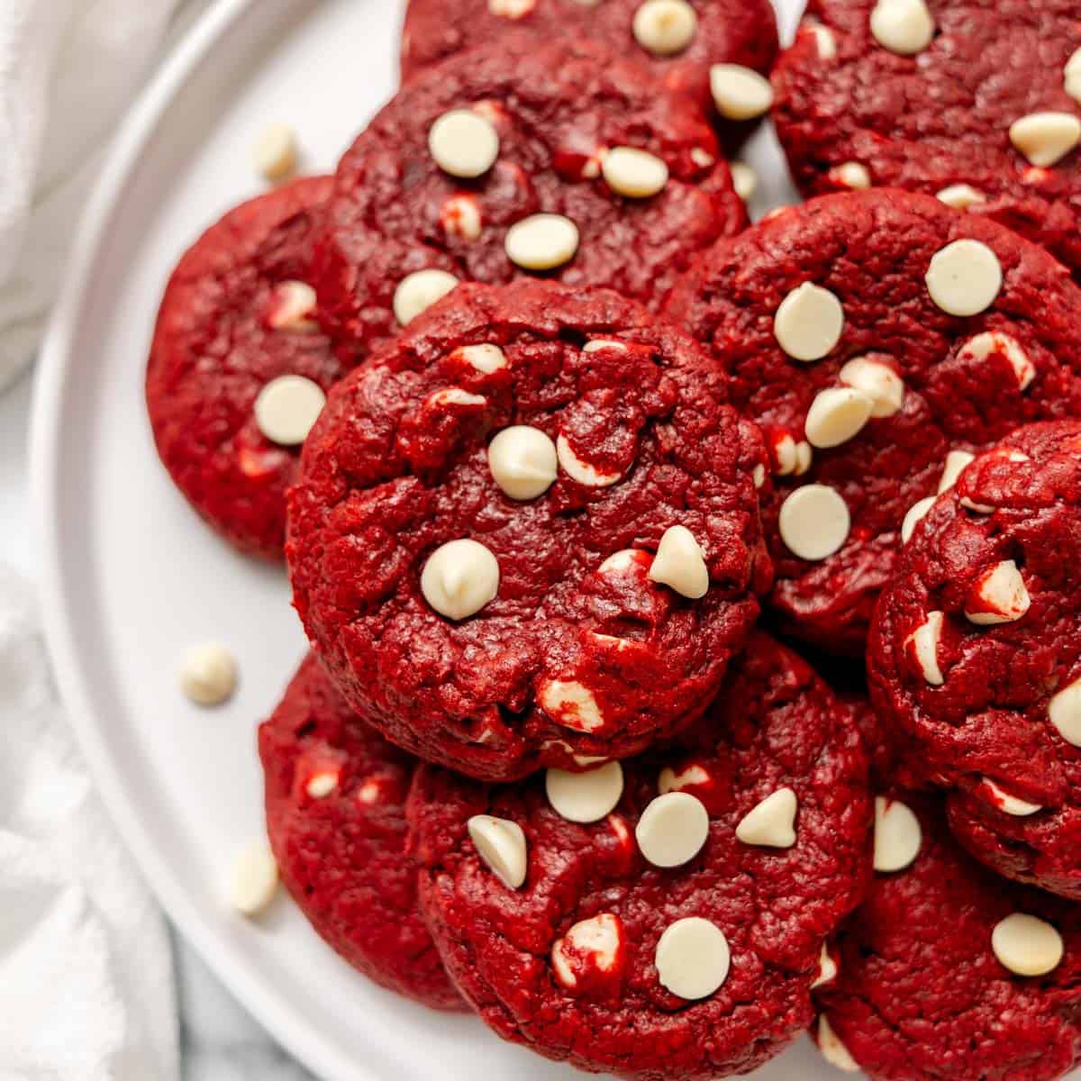 A pile of red velvet cookies studded with white chocolate chips on a white plate.