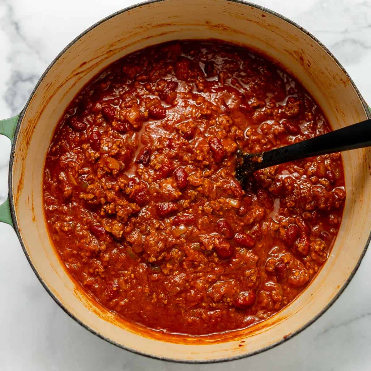 Large pot filled with chili made with ground meat, beans, and tomato sauce, with a black ladle resting inside.
