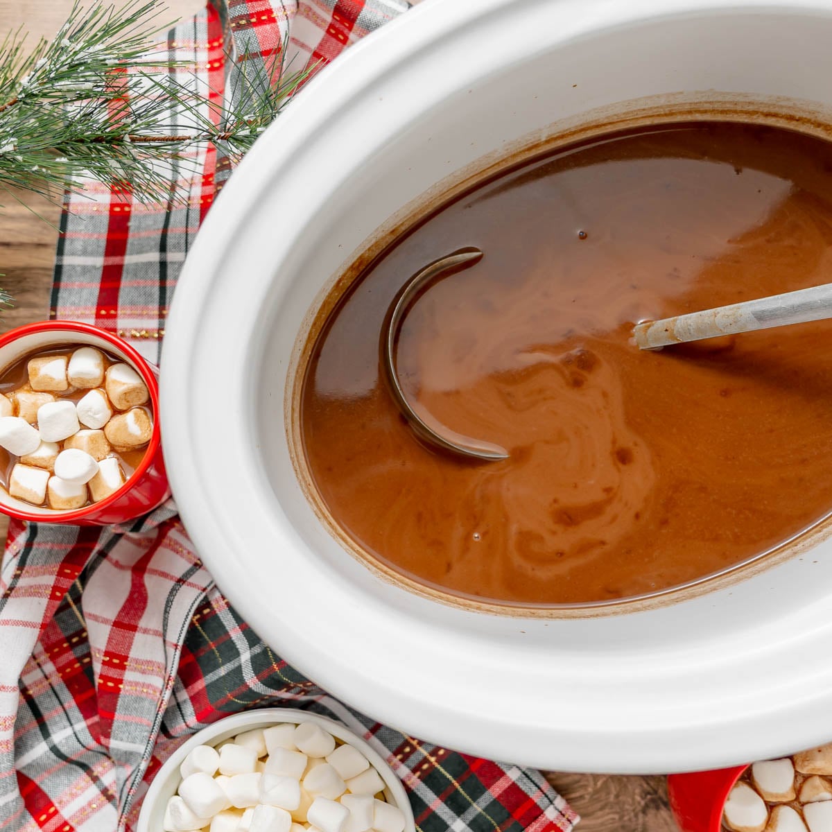 A slow cooker filled with hot cocoa and a ladle, with mugs and a bowl of marshmallows nearby on a plaid cloth.