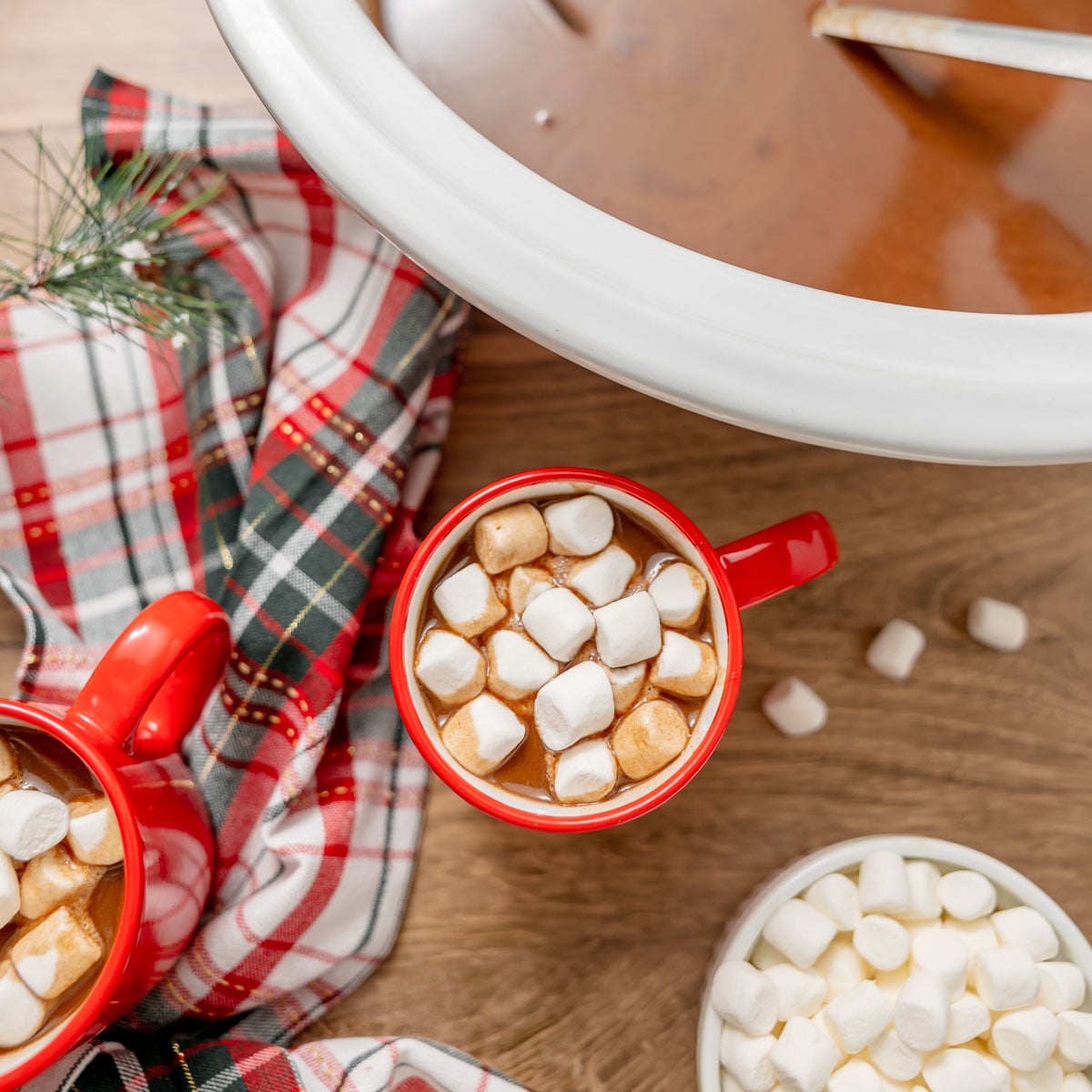 Two red mugs filled with hot chocolate and marshmallows, with a large Crockpot of hot chocolate and a small bowl of marshmallows nearby on a wooden table.