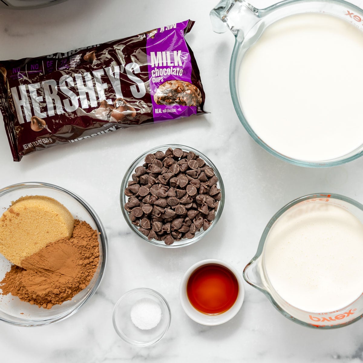 A package of Hershey’s milk chocolate chips with bowls of brown sugar, cocoa powder, loose chocolate chips, a small portion of salt, a splash of vanilla extract, and two pitchers of milk.