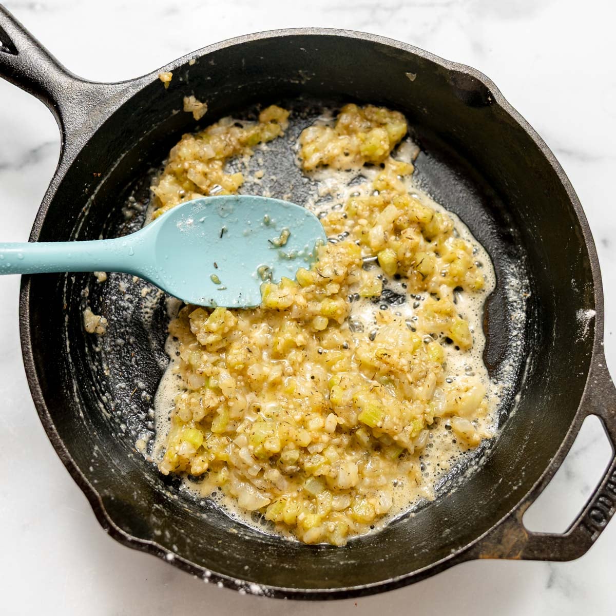 A light blue spatula stirs sautéed onions and celery coated in flour in a cast-iron skillet.