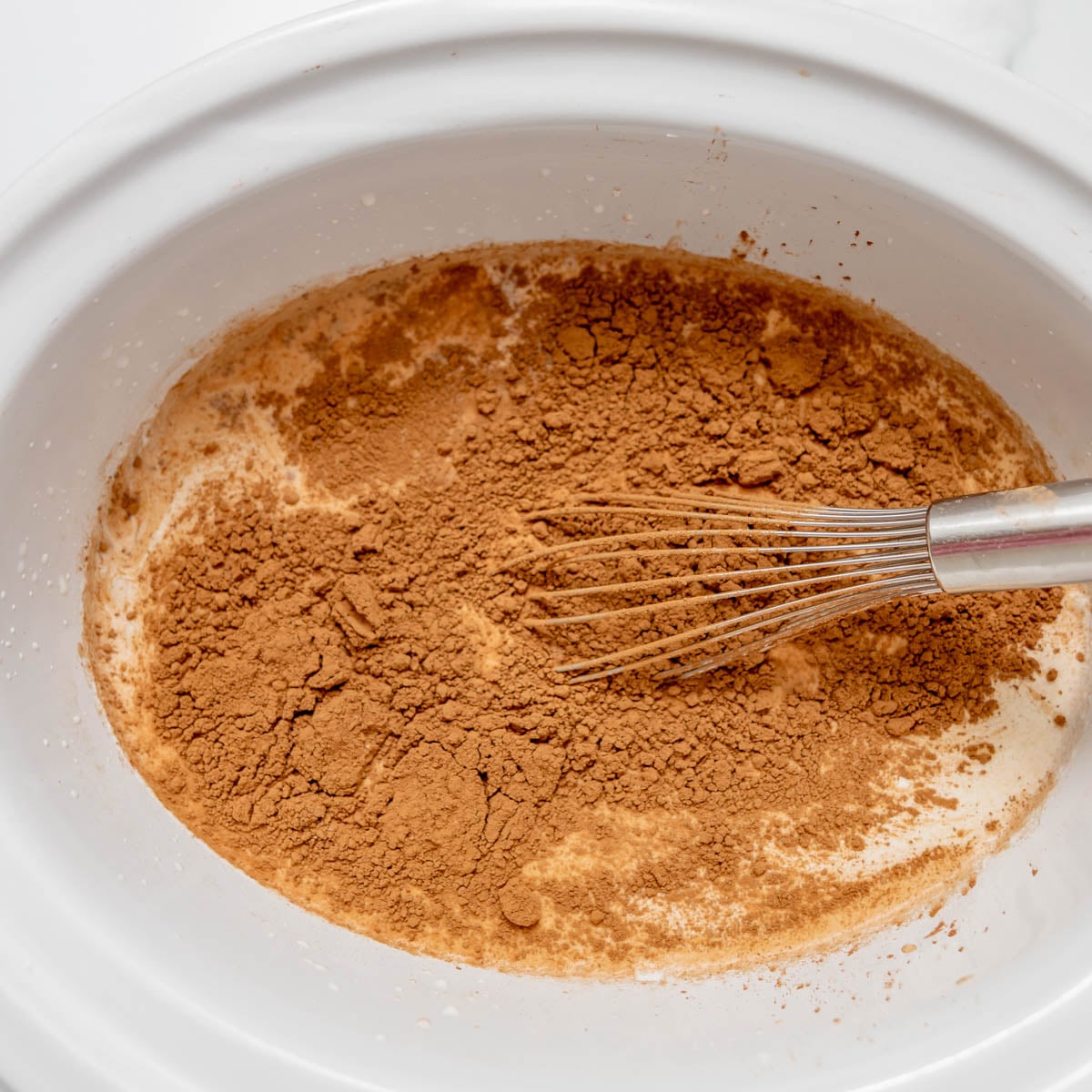 A whisk stirring cocoa powder into liquid in a slow cooker insert.