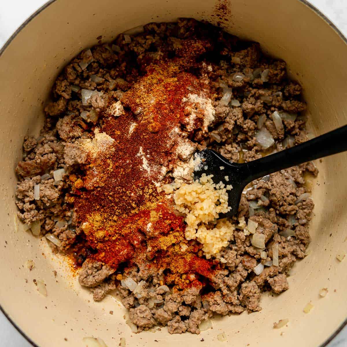 Ground beef and chopped onions cooking in a pot with minced garlic and various spices on top, being stirred with a black spatula.