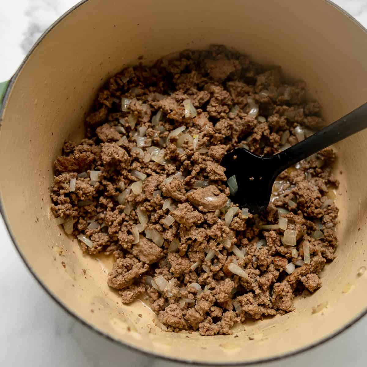 A pot filled with cooked ground beef and chopped onions being stirred with a black utensil.