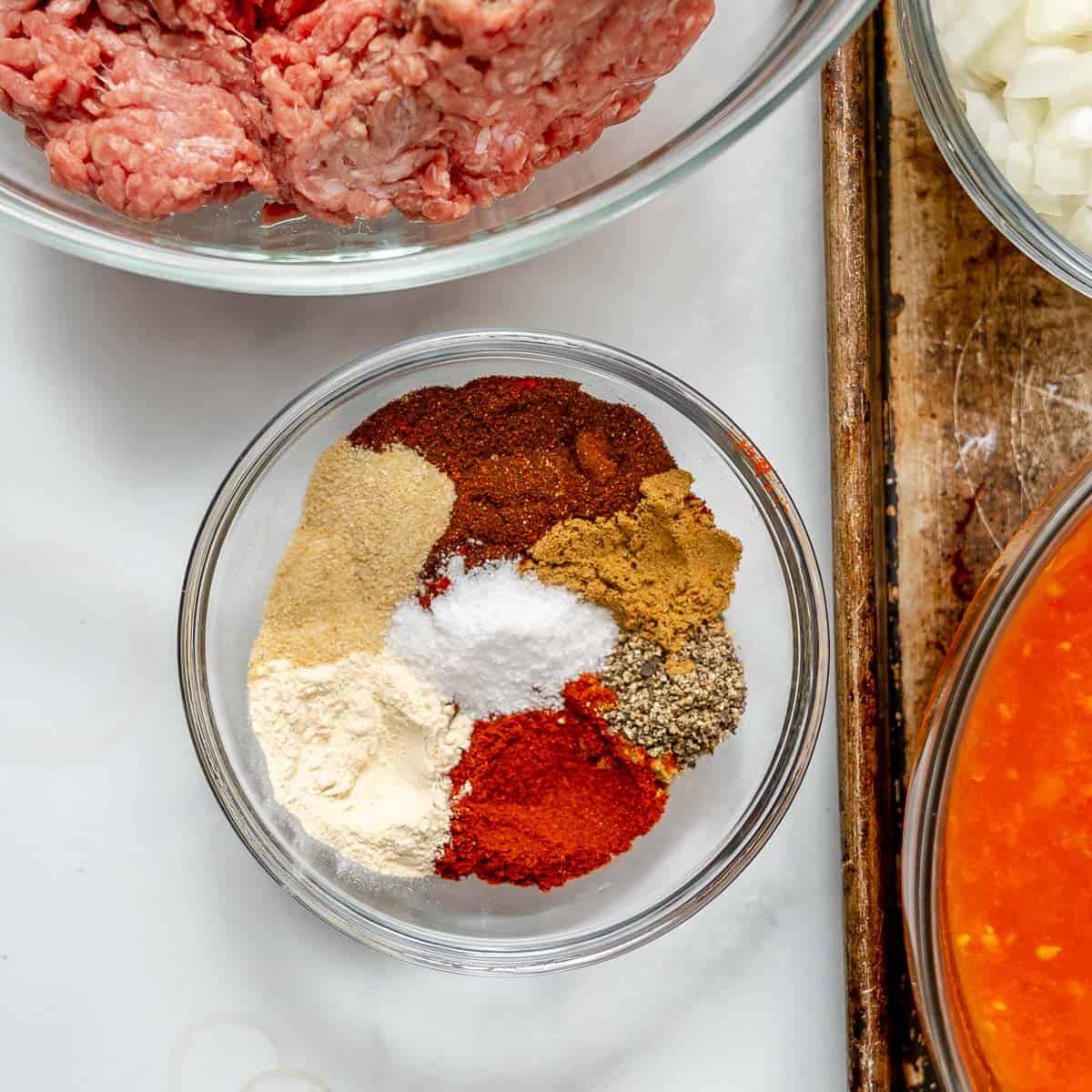 A glass bowl contains various ground spices arranged in sections. Nearby are bowls of raw ground meat, chopped onions, and a red-orange sauce.