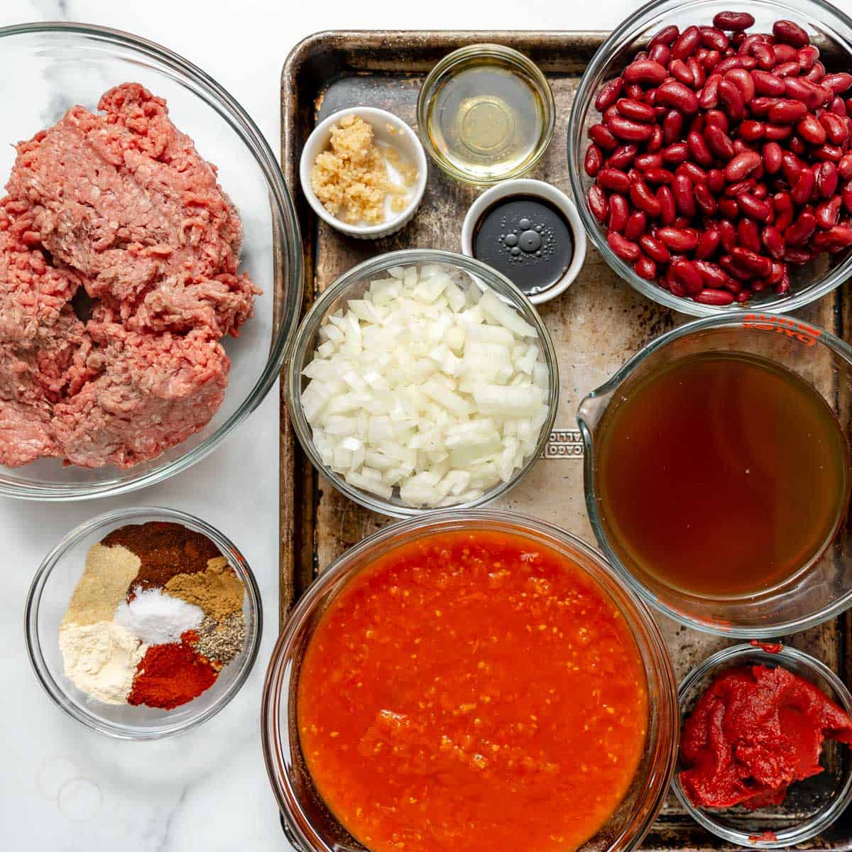Raw ground beef, chopped onions, minced garlic, red kidney beans, tomato sauce, tomato paste, beef broth, olive oil, soy sauce, and assorted spices arranged on a tray.