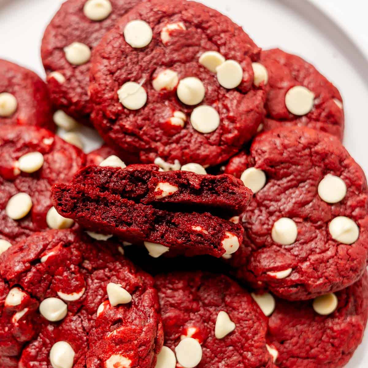 A pile of red velvet cookies studded with white chocolate chips, one broken in half to show the soft interior.
