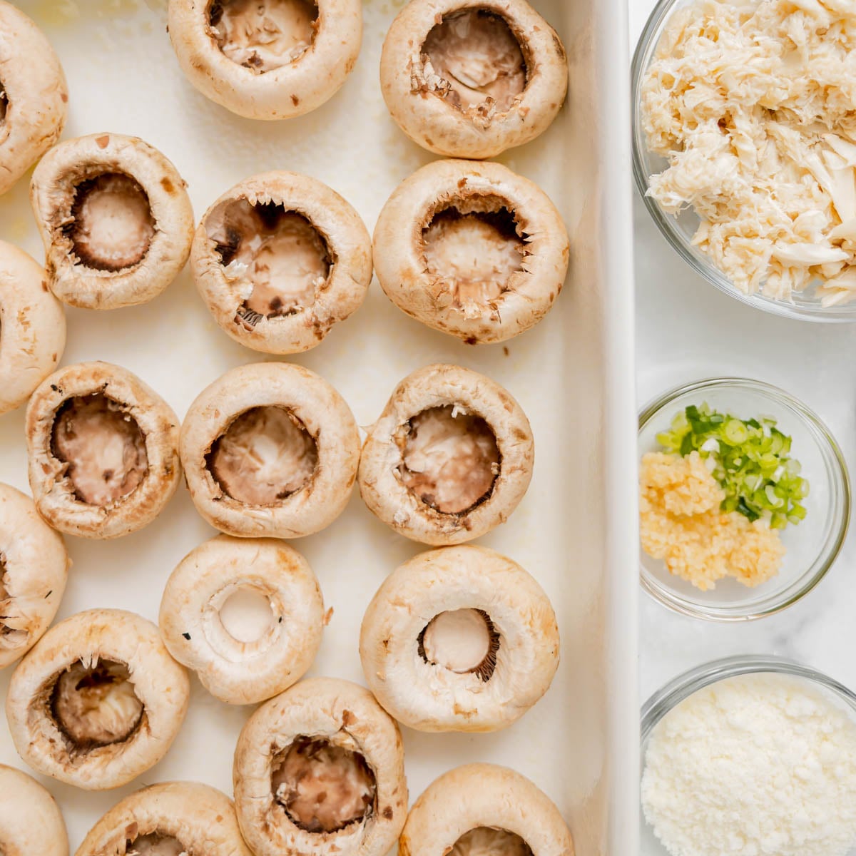 A baking dish filled with raw, hollowed-out mushroom caps next to small glass bowls containing shredded crab meat, chopped green onions with garlic, and grated cheese.