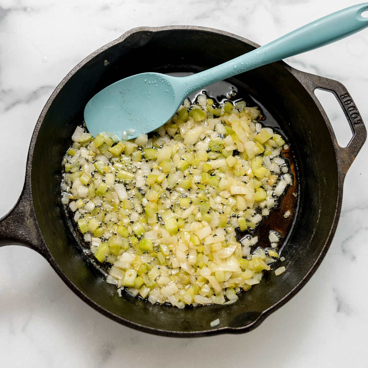Diced onions and celery sautéing in a cast-iron skillet with a blue silicone spatula.