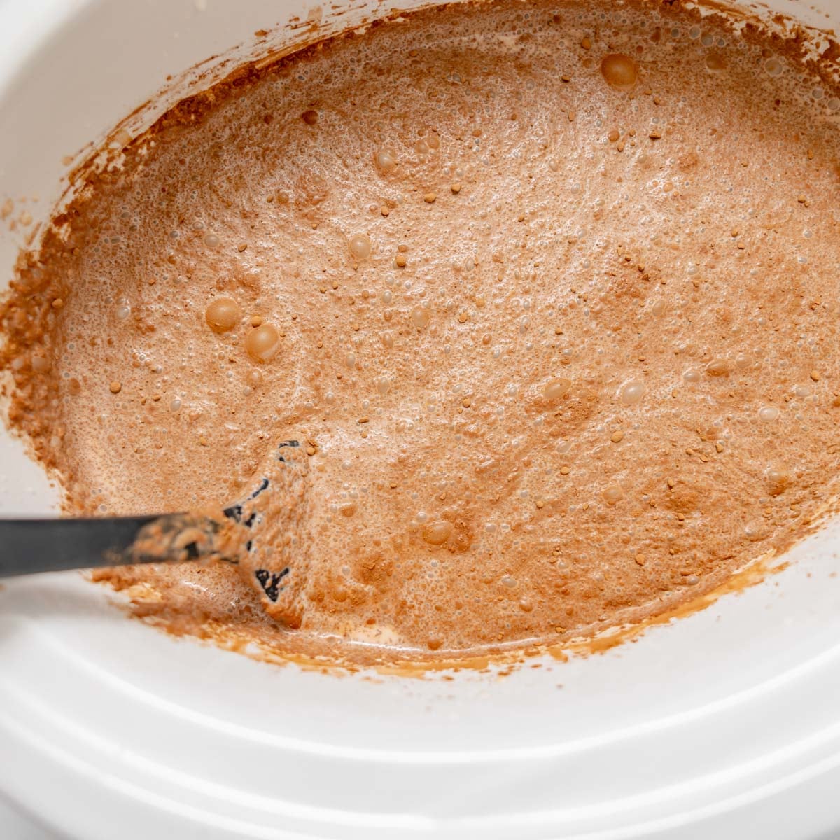 A spatula stirring a light brown, foamy liquid in a white slow cooker.