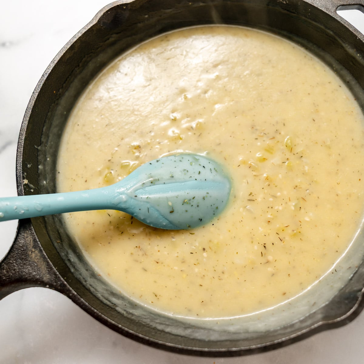 A light-colored, creamy sauce with visible herbs in a cast-iron skillet, being stirred with a blue silicone spoon.